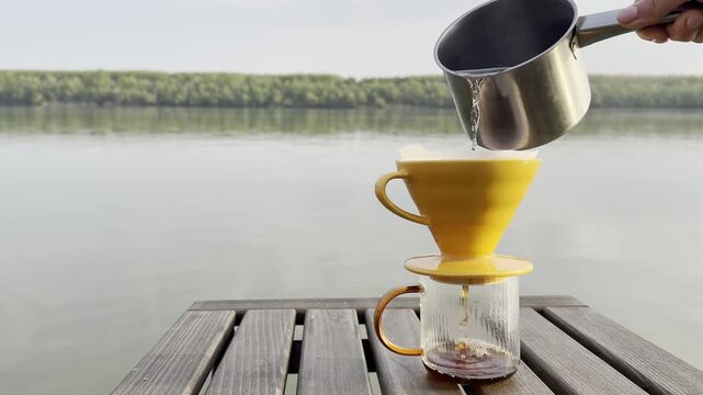 Close-up of hand pouring hot water into a pour over coffee dripper on a wooden table near a peaceful lake. Minimal outdoor coffee brewing scene with calm water, forest landscape, and soft natural