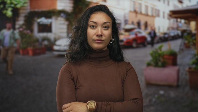 Woman with hand on chin and hoop earring on a cobblestone street beside potted planters and parked cars wearing brown turtleneck; thoughtful confidence.