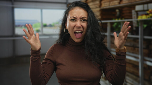 Woman with hands raised shouting inside building near shelves and window, wearing hoop earrings and brown turtleneck; anger dispute.