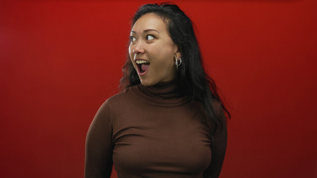 Woman with open mouth gasp and surprised gaze in studio on vivid red backdrop wearing brown turtleneck and hoop earrings; surprise joy energy.