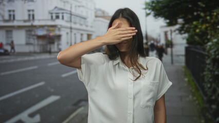 Fototapeta premium Young hispanic woman wearing glasses covers eyes with hand on street while smiling; playful confidence.