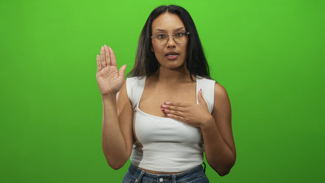 Woman with glasses in white tanktop raising right hand and placing left hand over chest in studio greenscreen, speaking as if taking an oath; commitment solemn.