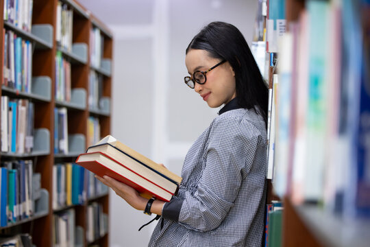 Young Asian woman studies in quiet library. Books line tall shelves behind. she holds  book, eyes focused. Serene atmosphere, calm concentration. Ideal for educational or study-themed projects