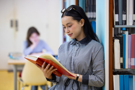 Young Asian woman studies in quiet library. Books line tall shelves behind. she holds  book, eyes focused. Serene atmosphere, calm concentration. Ideal for educational or study-themed projects