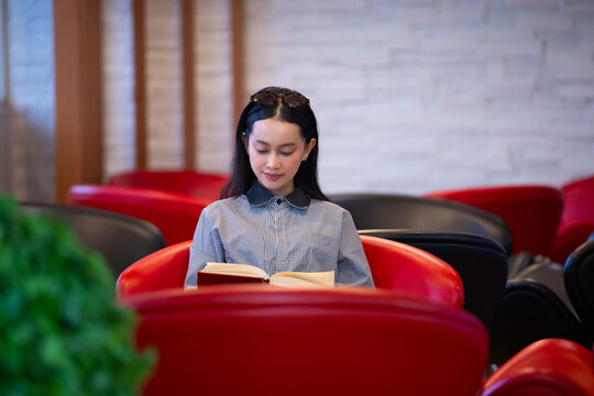 Young Asian woman studies reading book anf Siting on red chair in quiet library. Books line tall shelves behind. she holds book, calm concentration. Ideal for educational or study-themed projects