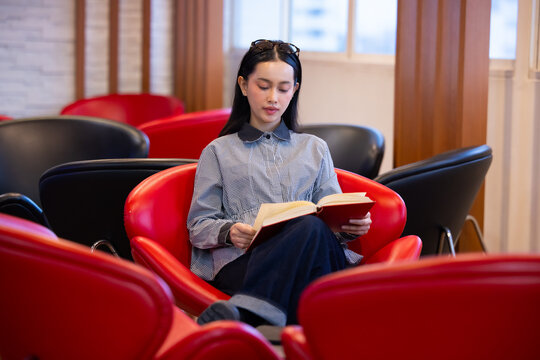 Young Asian woman studies reading book anf Siting on red chair in quiet library. Books line tall shelves behind. she holds book, calm concentration. Ideal for educational or study-themed projects