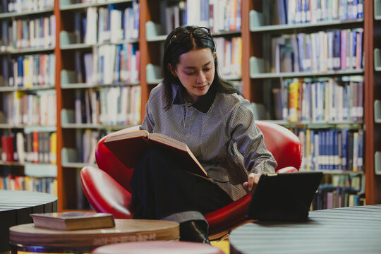 Young Asian woman studies reading book anf Siting on red chair in quiet library. Books line tall shelves behind. she holds book, calm concentration. Ideal for educational or study-themed projects