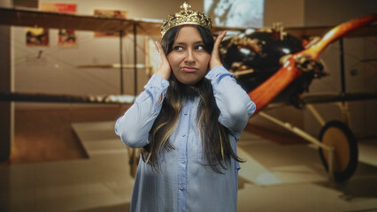 Hispanic woman wearing a gold crown covers her ears with both hands in a museum building beside a vintage biplane exhibit  annoyance. © Krakenimages.com