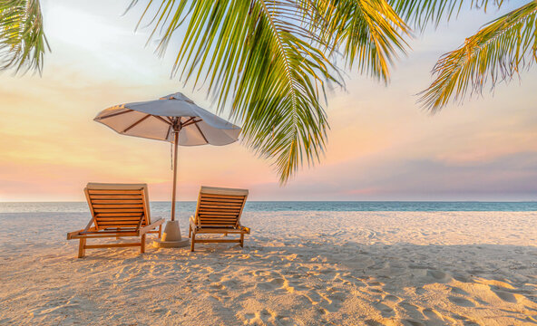 Wide panoramic view of beautiful tropical sunset scenery with two sun beds and umbrella under palm tree on a tranquil beach for inspirational beach resort hotel background and luxury wall art concept