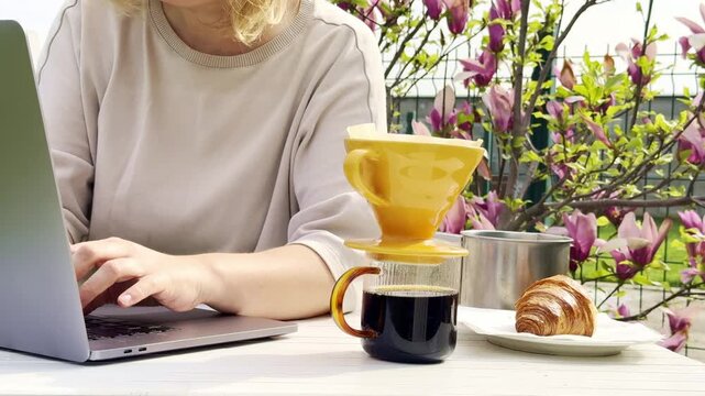Cozy outdoor remote work setup with freshly brewed pour over coffee, laptop, and croissant on a sunny spring morning. Lifestyle scene featuring blooming magnolia flowers and a relaxed home office 
