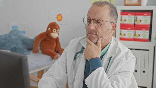 Man doctor, middle age, wearing glasses and stethoscope, hand on chin at computer in pediatric clinic with stuffed monkey on exam couch; thoughtful concern.