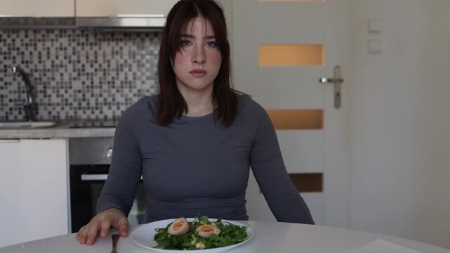 Young woman stretches measuring tape toward camera while eating healthy salad symbolizing pressure to stay thin and obsession with body measurements