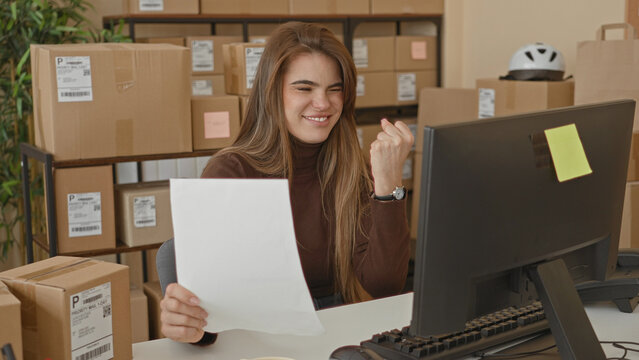 Woman holding a document and checking parcel labels at a computer desk amid stacked packages in a packing building; small business satisfaction.