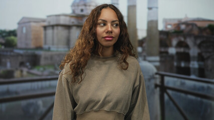 Obraz premium Woman looking sideways with a subtle smile, wearing a loose sweatshirt on a building terrace overlooking roman ruins and columns; serenity.