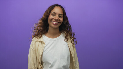 Fototapeta premium Woman smiling with eyes closed showing teeth and loose curly hair, wearing white t shirt and beige overshirt in a purple studio; contentment.