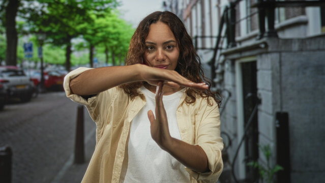 Young woman making a time out gesture with hands on a city street lined with parked cars and trees; stop boundary determination.