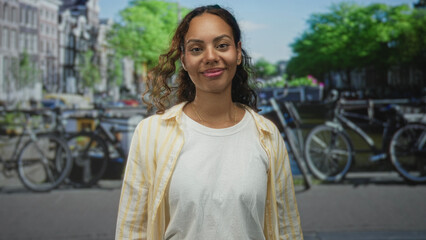 Fototapeta premium Young african american woman smiling with hand to chest on amsterdam street with parked bicycles and canal behind her; confidence.