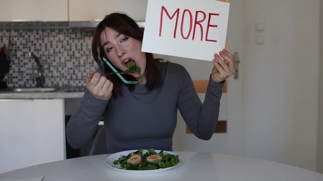 Young employee eats lunch during nonstop work calls while holding sign with word more symbolizing modern obsession with productivity success and constant pressure