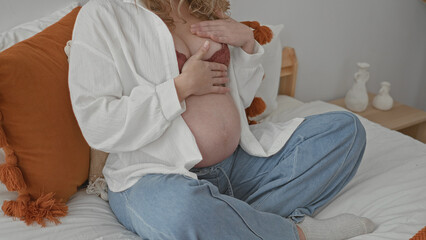 Pregnant woman sitting on bed touching bare belly, hand on chest with open shirt and visible bra in...