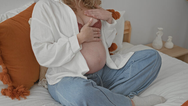 Pregnant woman sitting on bed touching bare belly, hand on chest with open shirt and visible bra in studio setting; motherhood bonding serenity.