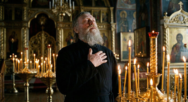 Senior Orthodox monk with closed eyes praying with hand on heart in candlelit church before iconostasis.