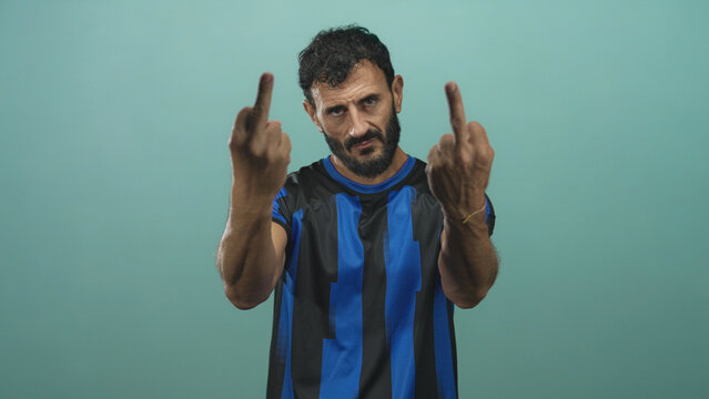 Man in blue and black soccer jersey shows middle finger to camera in studio, bearded player with stern face, extended arm and provocative gesture; defiance protest.