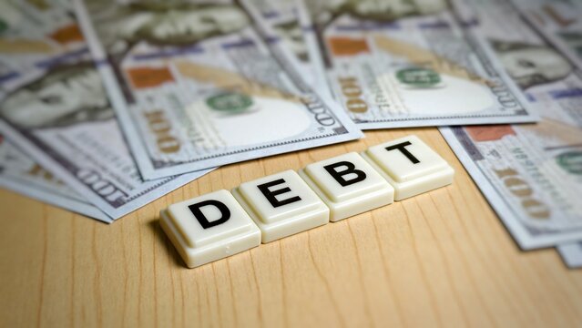 The word "DEBT" is spelled with tile blocks on a wood surface, surrounded by scattered US $100 bills, illustrating financial obligation, borrowing, and the concept of monetary deficit.