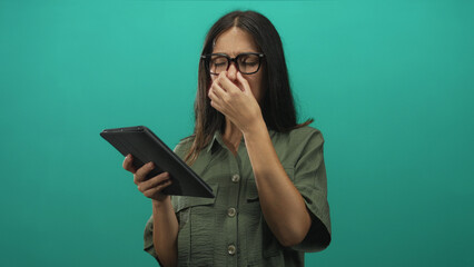 Young hispanic woman pinches her nose holding a tablet in a teal studio backdrop wearing glasses and olive shirt  disgust. © Krakenimages.com