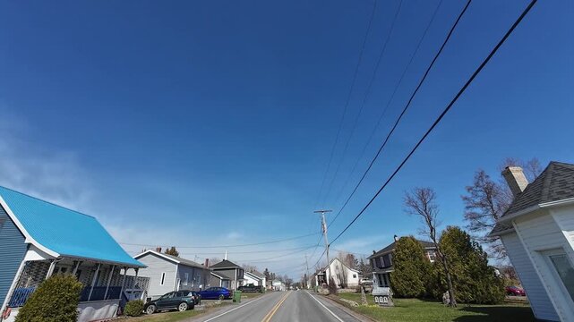 4K dashcam footage traveling along Route 297 through Saint-Damase village with church steeple, residential homes and scenic Matapedia Valley landscapes under blue skies in Quebec, Canada.