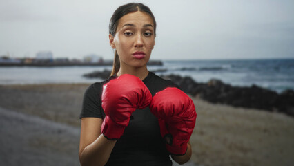 Fototapeta premium Woman wearing red boxing gloves holds fists up on a beach with a distant building and rocky shoreline; determination grit.