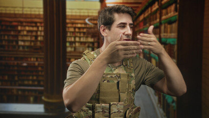 Man soldier wearing camouflage vest and dogtags blows kiss with right hand to mouth near bookshelves inside building library  affection memory longing. © Krakenimages.com
