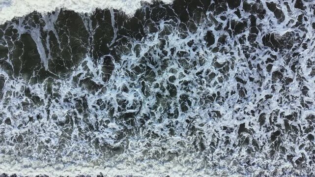 Waves roll in from the ocean, hitting the shore and forming foamy patterns on the sand. The bright sunlight reflects off the water, creating a dynamic scene.