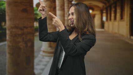 Fototapeta premium Woman points both index fingers upward while smiling in a cloister corridor, showing fingers and playful gesture; joy.