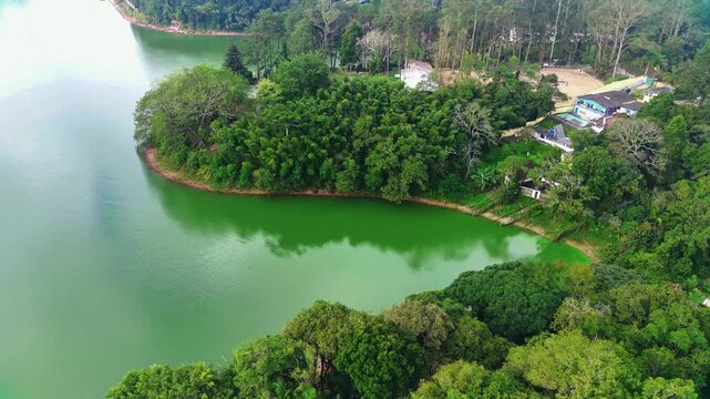 Vista a&eacute;rea de lago verde cercado por floresta e casas &agrave; beira d&rsquo;&aacute;gua.