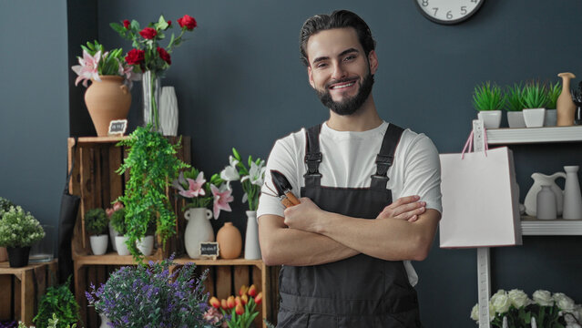 Man florist holding pruners with crossed arms among vases and flowers in building shop interior, smiling at camera; friendly confidence.