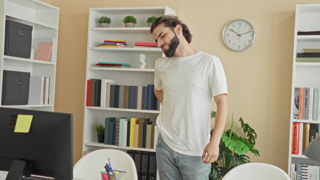 Young hispanic man touching lower back by computer monitor in an office building, wincing from backache; discomfort posture strain.