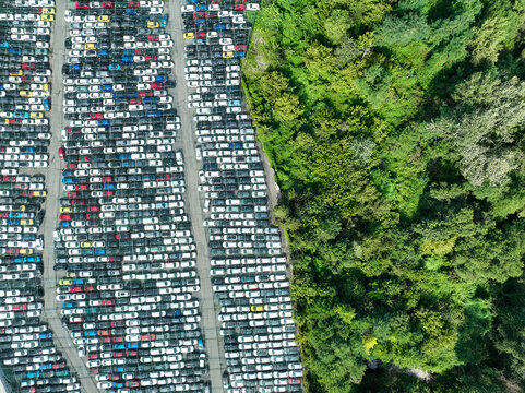Aerial view of a large parking lot filled with rows of new cars next to a lush green forest in Turin, Piedmont, Italy.