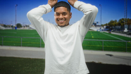 Man with beard and hands on head smiling showing teeth in white long sleeve shirt at a stadium building, soccer field visible  joy casual. © Krakenimages.com
