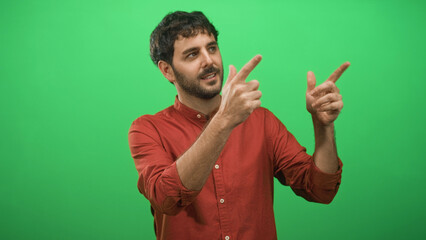 Fototapeta premium Man with beard points fingers with both hands toward right in studio green screen; playful presentation.