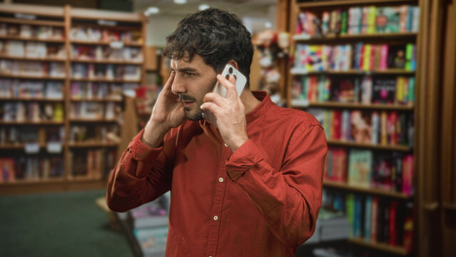 Hispanic man holding smartphone to ear and cupping head with hand in a bookstore building; frustration urgency.