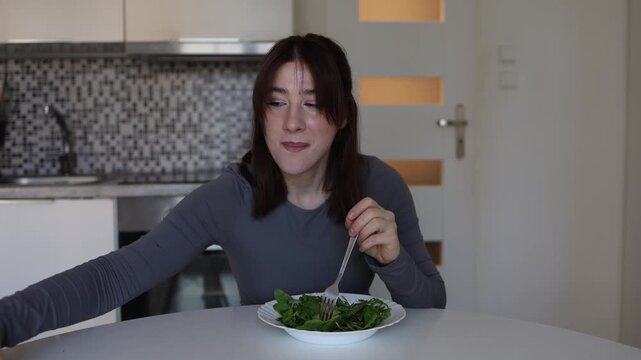 Young woman trying to eat healthy salad loses self control and reaches for sweet chocolate craving sugar while sitting alone in modern kitchen at home