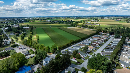 Obraz premium A view of a small town surrounded by green fields and farm areas. The houses are in neat rows, while the fields stretch across the landscape. The sky is partly cloudy and blue.