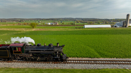 A steam train moves along tracks surrounded by green fields and farmland. The sun shines on the landscape, showing barns and silos in the distance. The scene captures rural life and train travel. © Greg Kelton