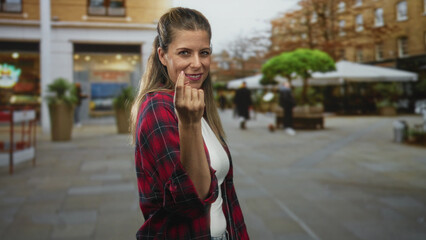 Fototapeta premium Woman beckons with finger on street by outdoor cafe and planters, wearing red plaid shirt and white top, smiling at camera; playful invitation.