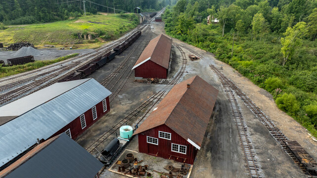 The scene shows multiple red storage buildings near train tracks. The area is green with trees and a slight incline in the background. Equipment and materials are scattered around the site.