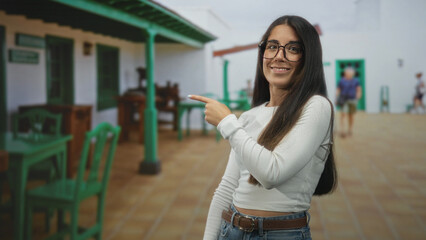 Fototapeta premium Young hispanic woman wearing glasses and denim points finger to green chair beside a white building with tiled courtyard and outdoor cafe tables; playful hush.