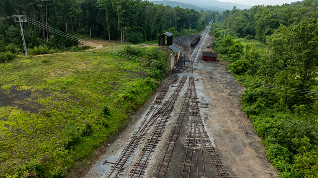Train tracks stretch into the distance through a green landscape. Abandoned buildings dot the area, surrounded by trees and hills. Power lines run alongside the tracks, connecting the scene.