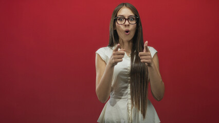 Fototapeta premium Young woman points fingers toward camera with hands visible in a red studio, surprised open mouth gesture; playful affection.