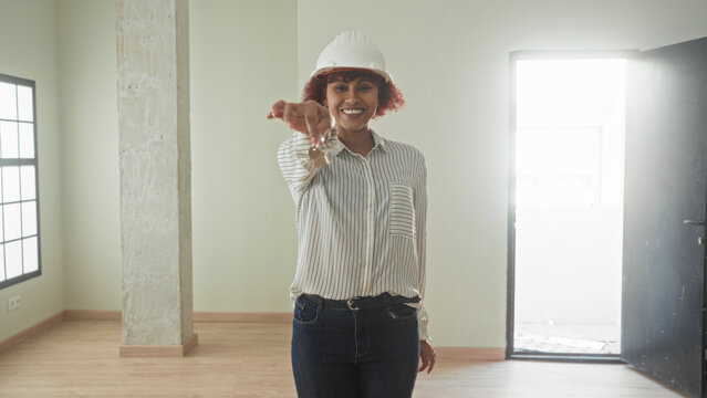Woman holding keys with outstretched hand in an empty building room, smiling while wearing hardhat and striped shirt as architect presenting access; homeowner pride.
