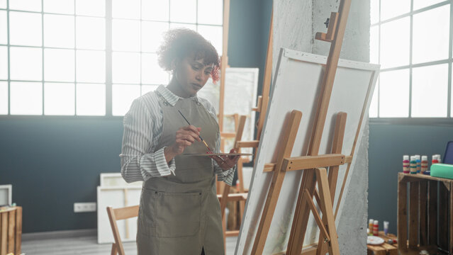 Woman painter paints canvas with brush and holds palette at wooden easel in art studio with large window light; creative focus.